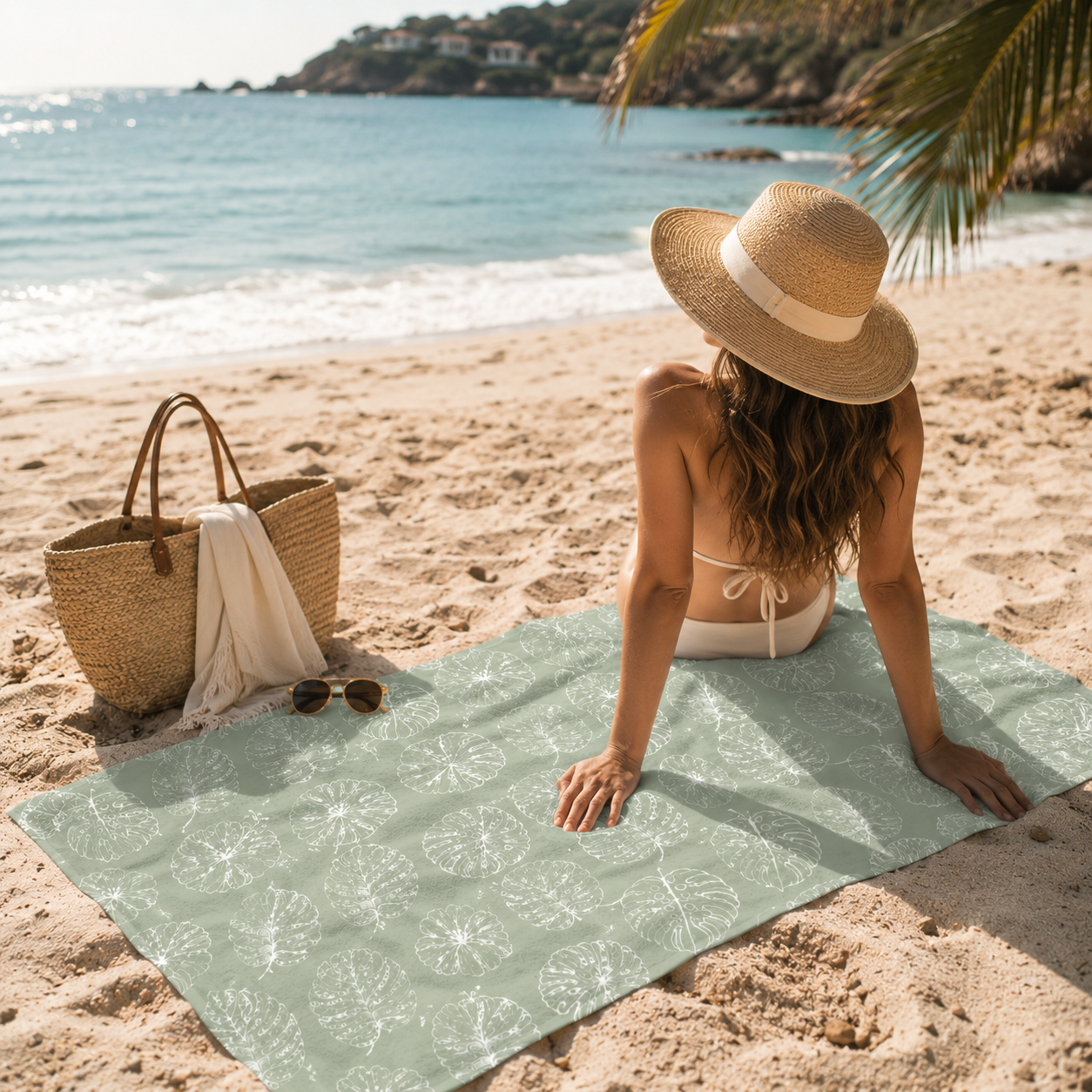 Woman sitting on a beach towel with a hat and bag, enjoying the view of the ocean.