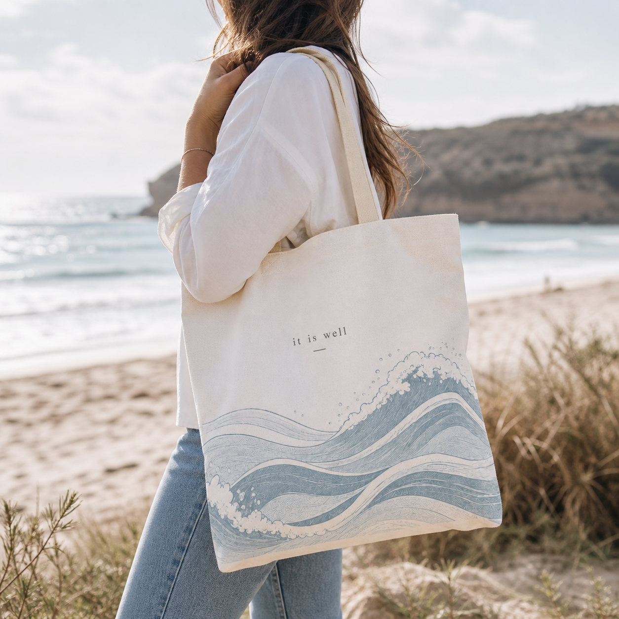Person holding a tote bag with wave design and 'it is well' text on a beach.
