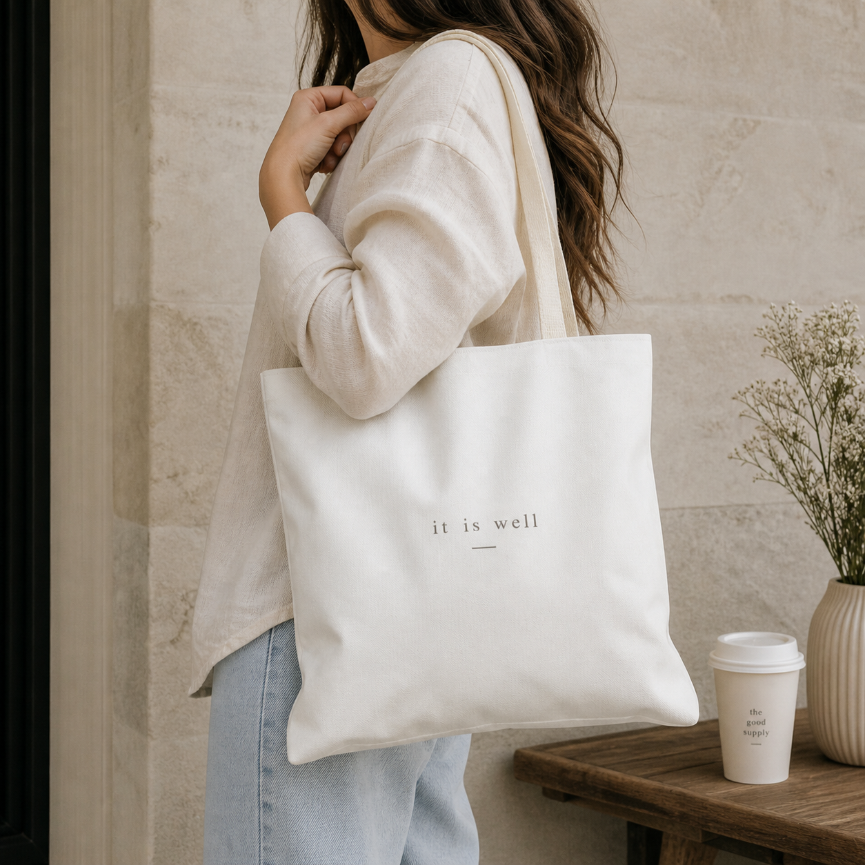Person holding a white tote bag with 'it is well' text, standing indoors.