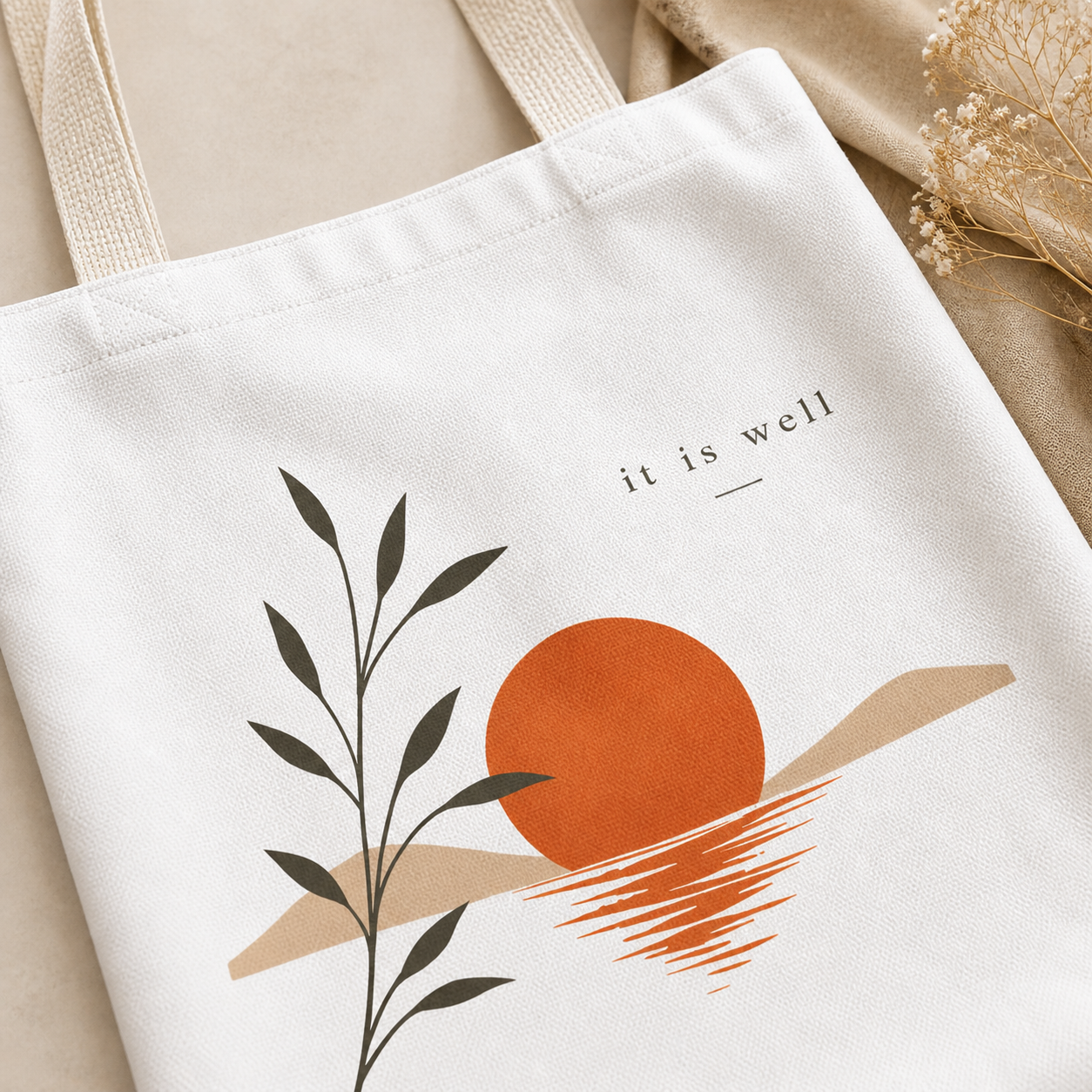 White tote bag with artistic design of a sun, leaves, and text 'it is well' on a beige background.