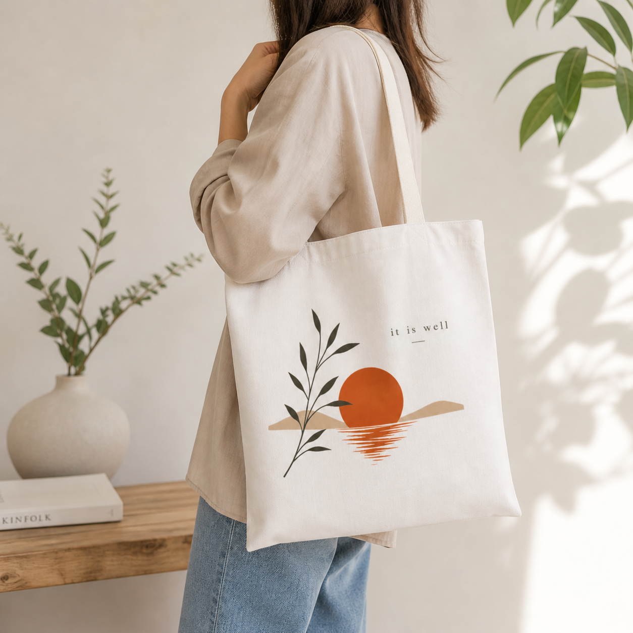 Person holding a tote bag with a design of a sun and leaves, text 'it is well', in a home setting.