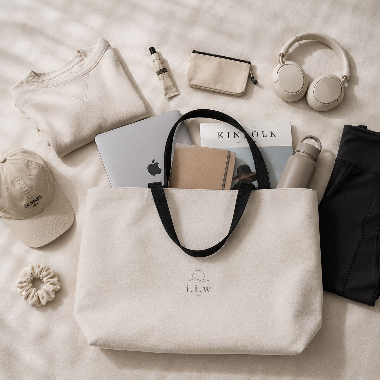 Beige tote bag with 'i.i.w' logo surrounded by various items on a light surface