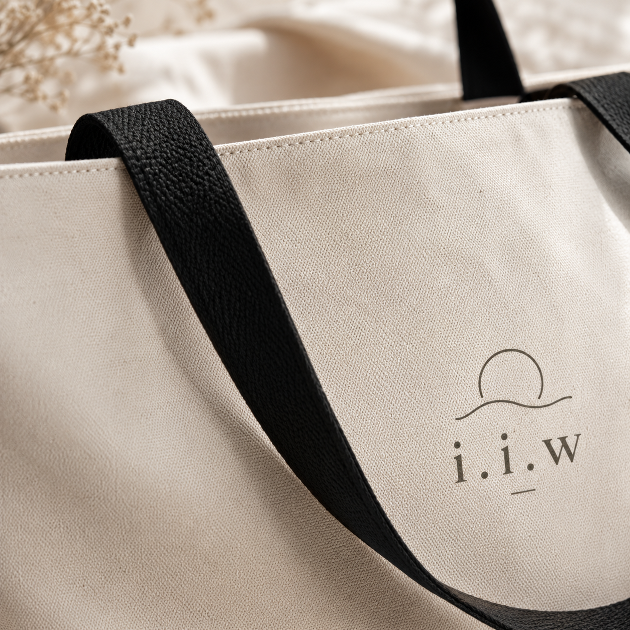 Beige tote bag with black handles and 'i.i.w' logo on a blurred background