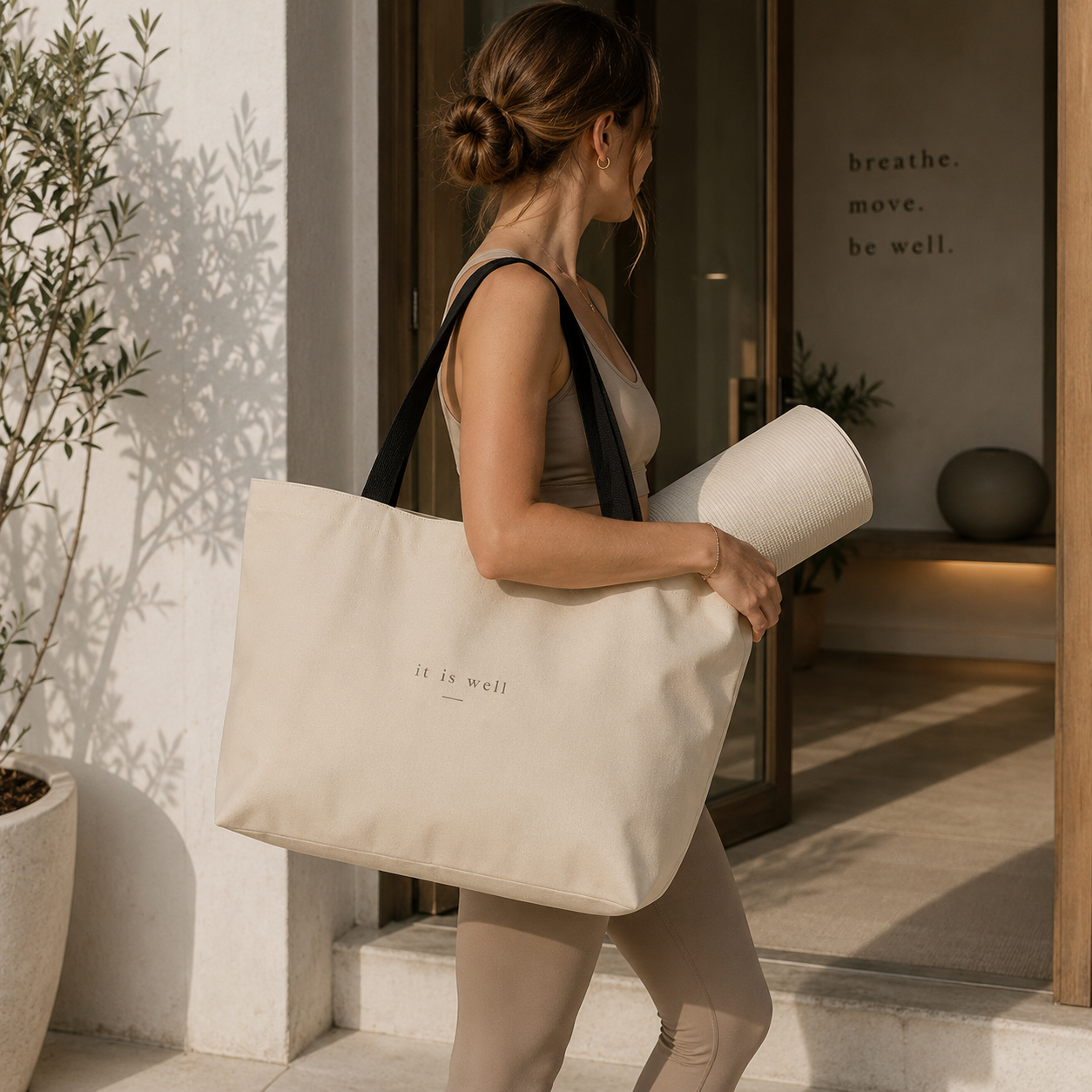 Woman holding a yoga mat and a tote bag with 'it is well' text, standing in front of a building with a plant.