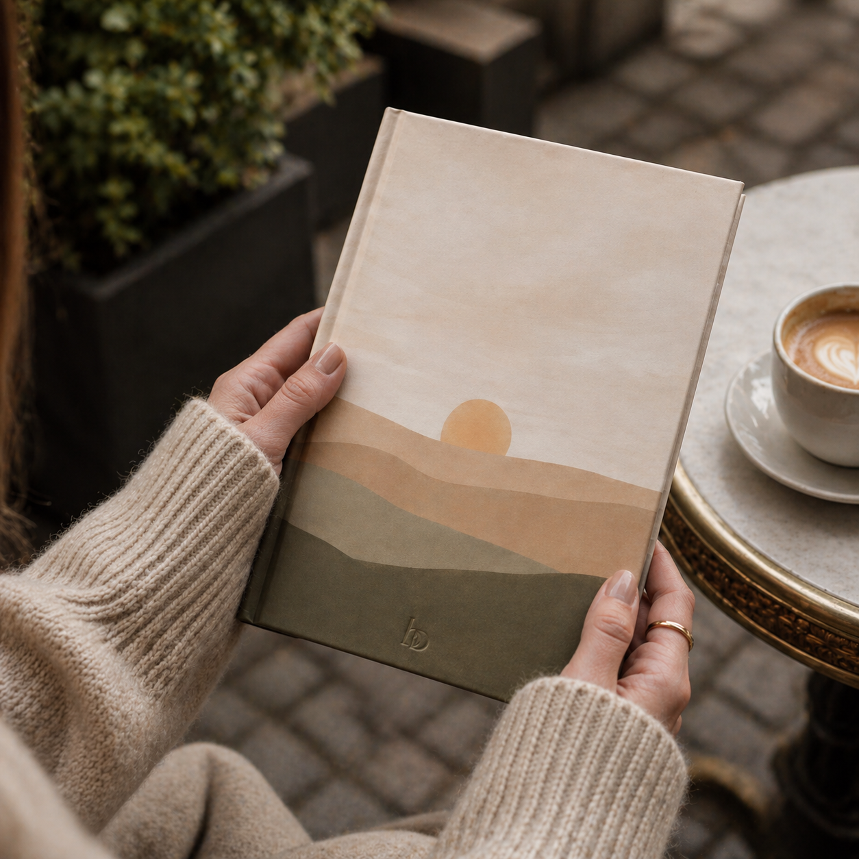 Person holding a notebook with a sunset design outdoors near a table with a coffee cup.