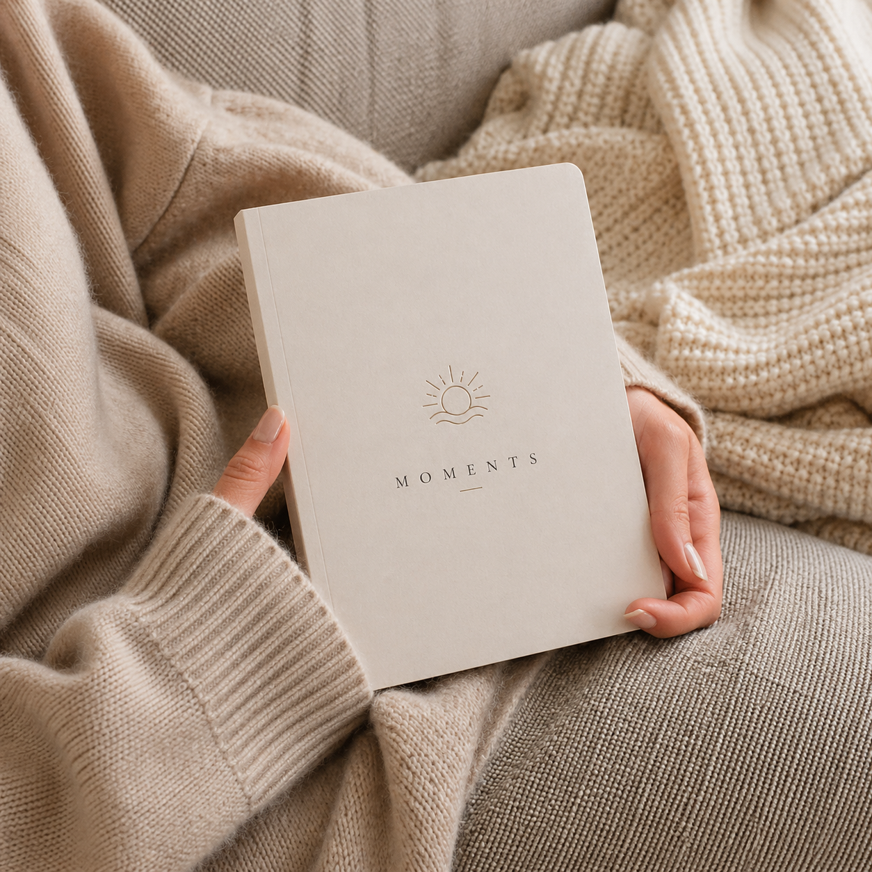 Person holding a beige notebook titled 'Moments' with a sun illustration, wearing a beige sweater.