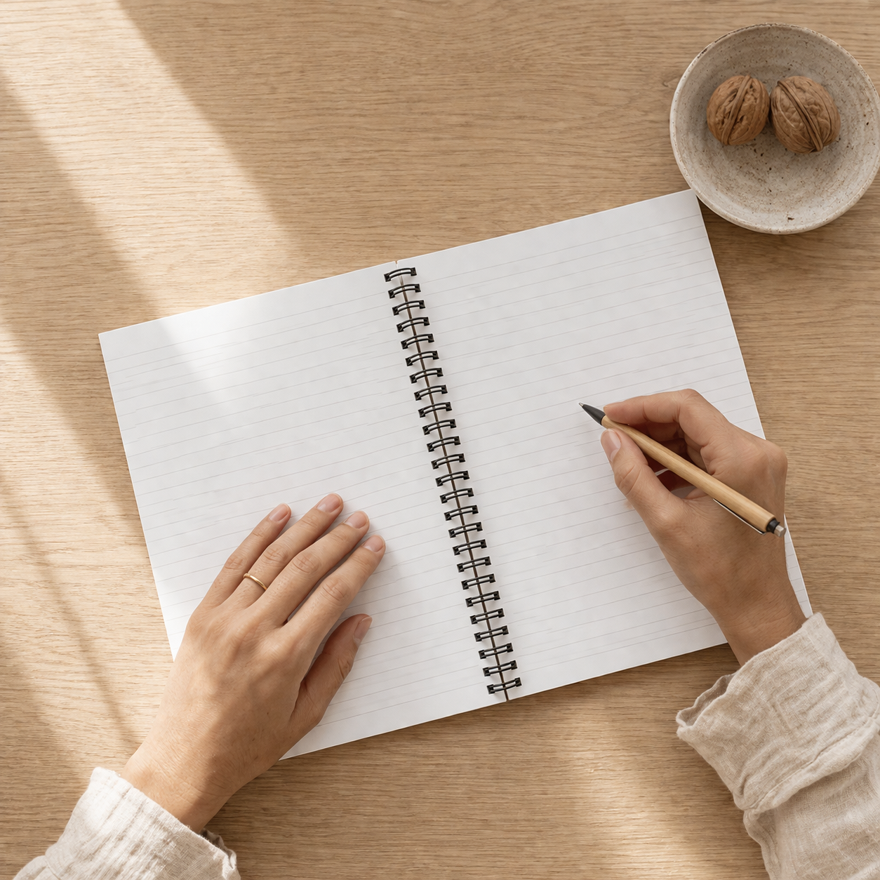 Person writing in a spiral notebook on a wooden table with a small bowl of nuts.