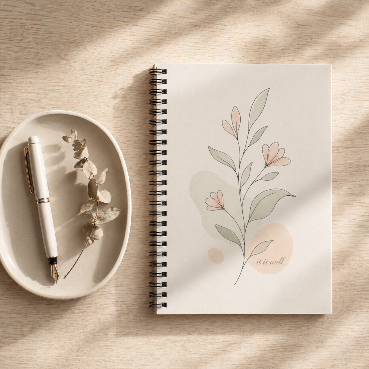 Spiral-bound notebook with floral design and 'it is well' text on a wooden surface with a pen and small dish.