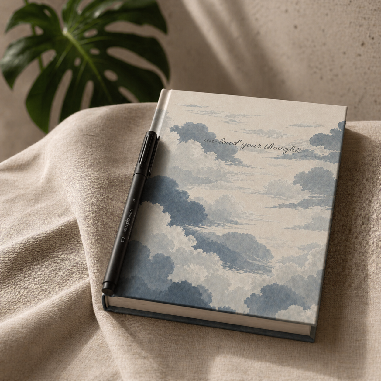 Notebook with cloud design and pen on beige fabric with plant in background