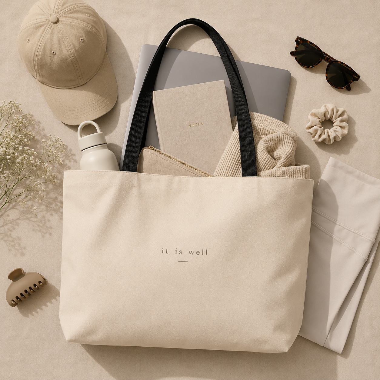 Beige tote bag with 'it is well' text, surrounded by a cap, notebook, sunglasses, and other items on a light background.